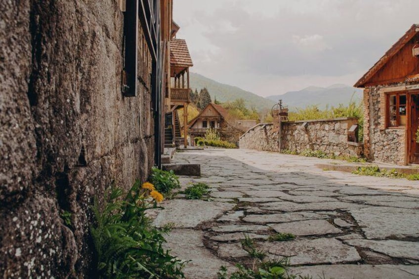 A stone-paved street with rustic buildings, a wooden fence, and mountains in the background. 5