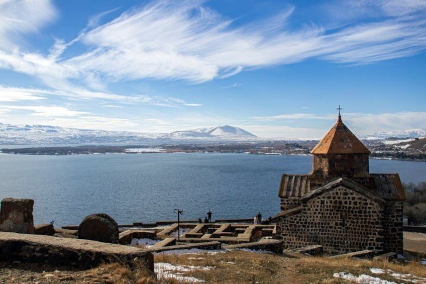 A historic church with a stone facade and a conical roof, overlooking a calm lake with a snow-capped mountain in the distance. 1