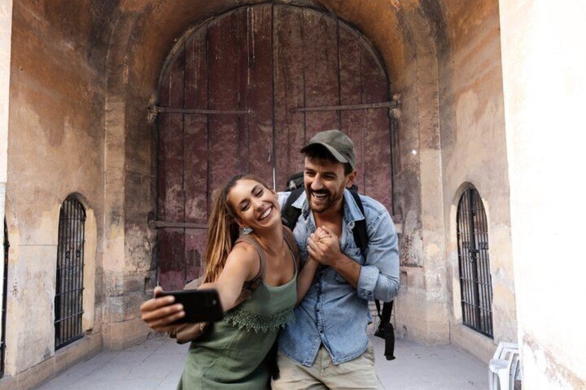 Zwei Personen machen ein Selfie vor einem alten Gebäude mit Rundbogenfenstern und einer großen Holztür. 1