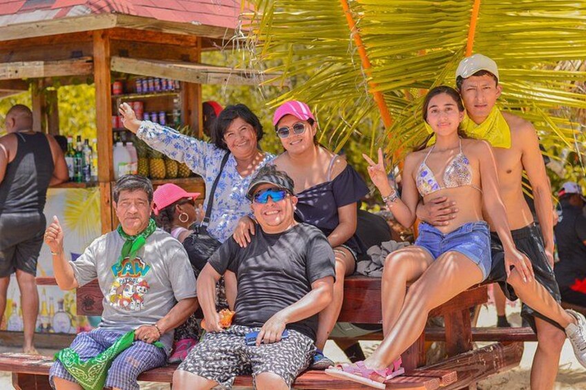 A group of people sitting on a wooden bench under a palm tree, with a beach and a small structure in the background. 4