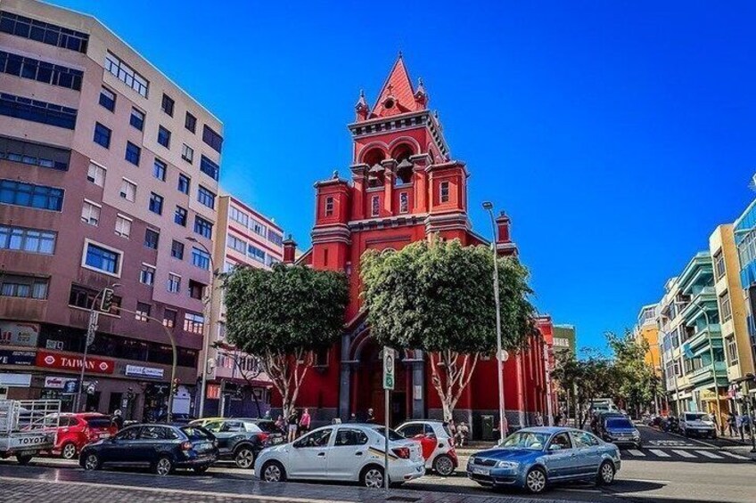A red church with a pointed roof, surrounded by modern buildings and parked cars. 5