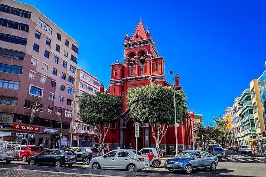 A red church with a pointed roof, surrounded by modern buildings and cars on a city street. 5