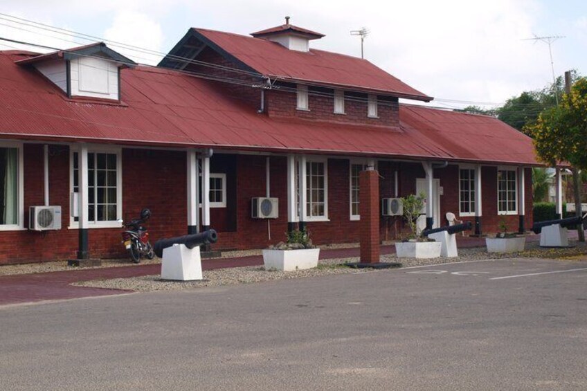 A red-roofed building with large windows and air conditioning units, surrounded by white planters and two cannons. 1
