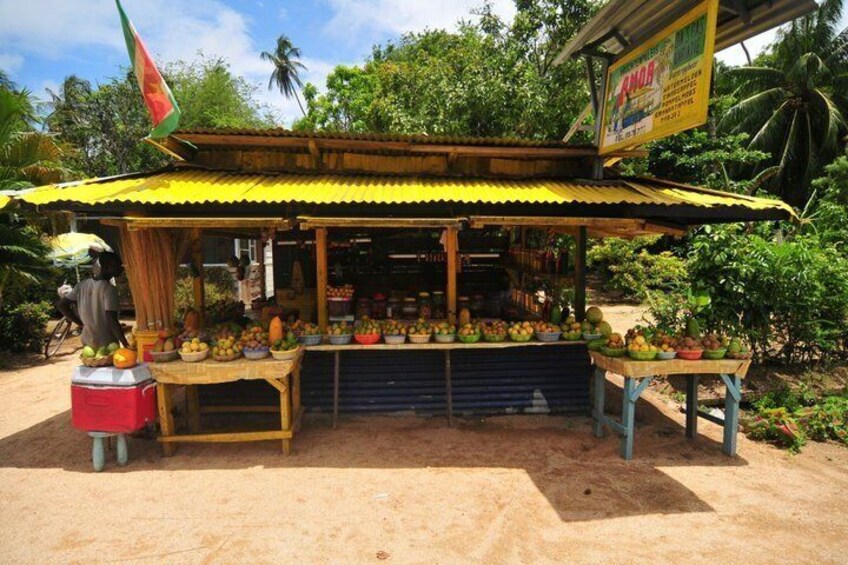 A fruit stand with a yellow roof, surrounded by greenery and palm trees. 3