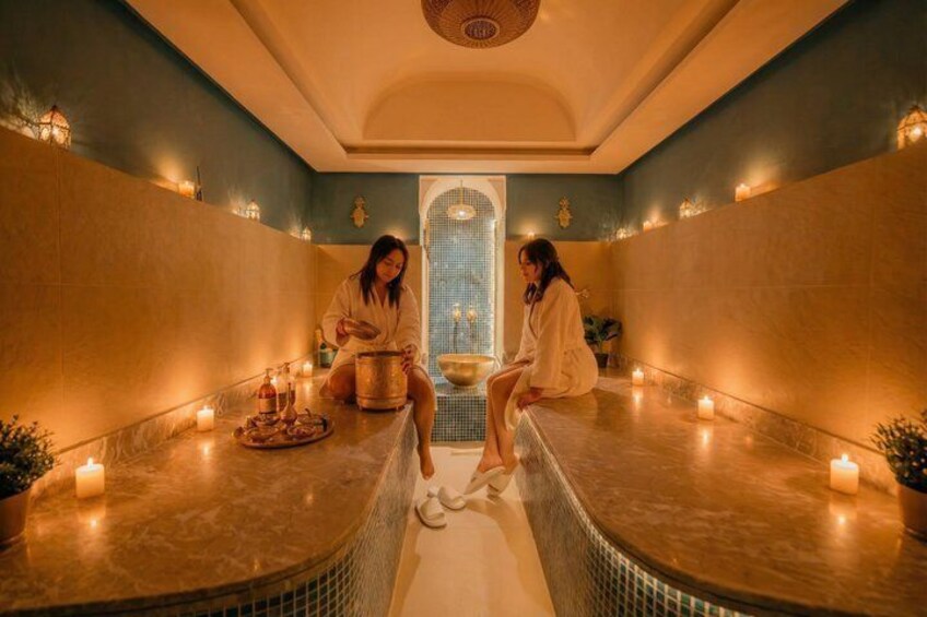Un spa où deux personnes sont assises sur des bancs en marbre, avec des bougies et une fontaine décorative. 1