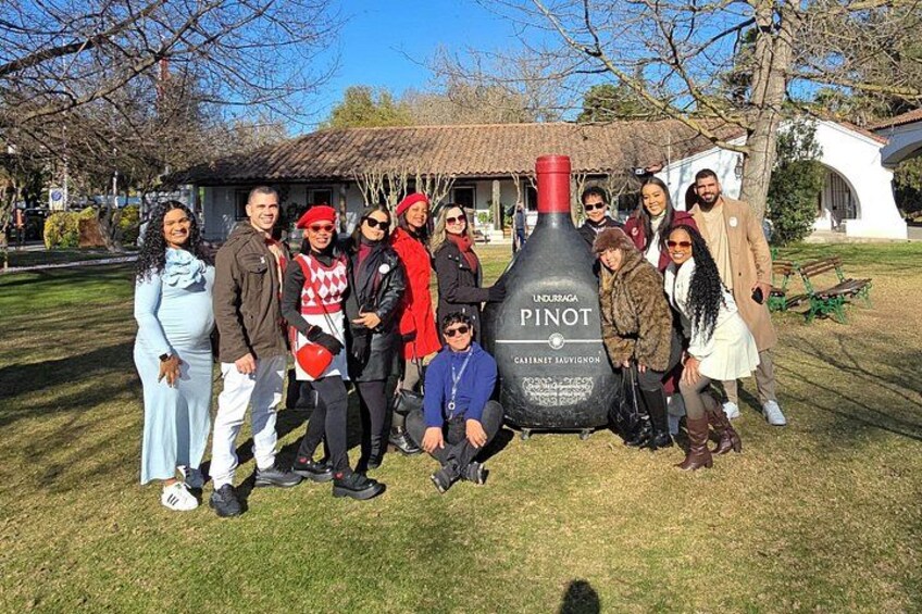 Un grupo de personas posando con una botella grande etiquetada como 'Pinot' en un área con césped y un edificio al fondo. 1
