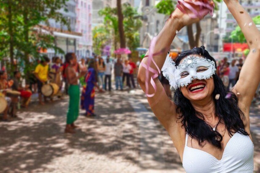 Una persona con un vestido blanco y los brazos levantados, posiblemente bailando, en un entorno al aire libre con árboles y edificios al fondo. 1
