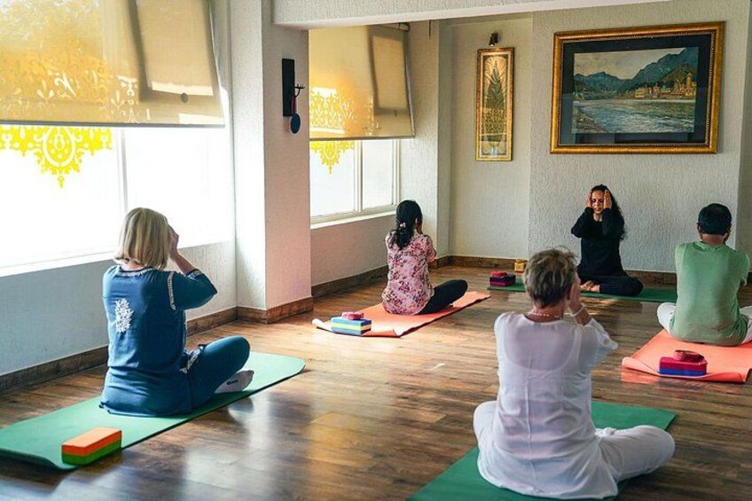 Un grupo de personas sentadas sobre esterillas de yoga en una habitación con suelos de madera y ventanas amplias. 1