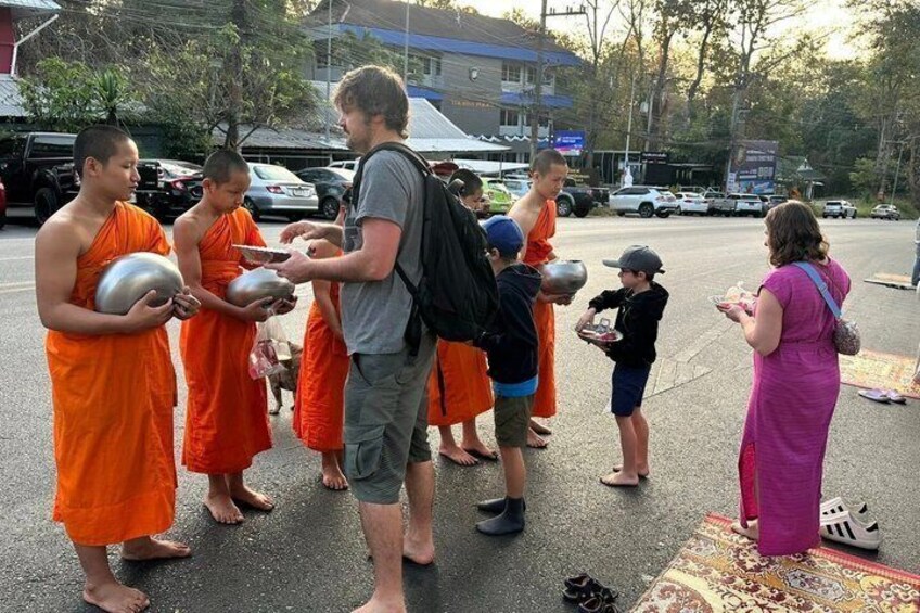 Monks in orange robes holding bowls, interacting with a man and a woman on a street. 2