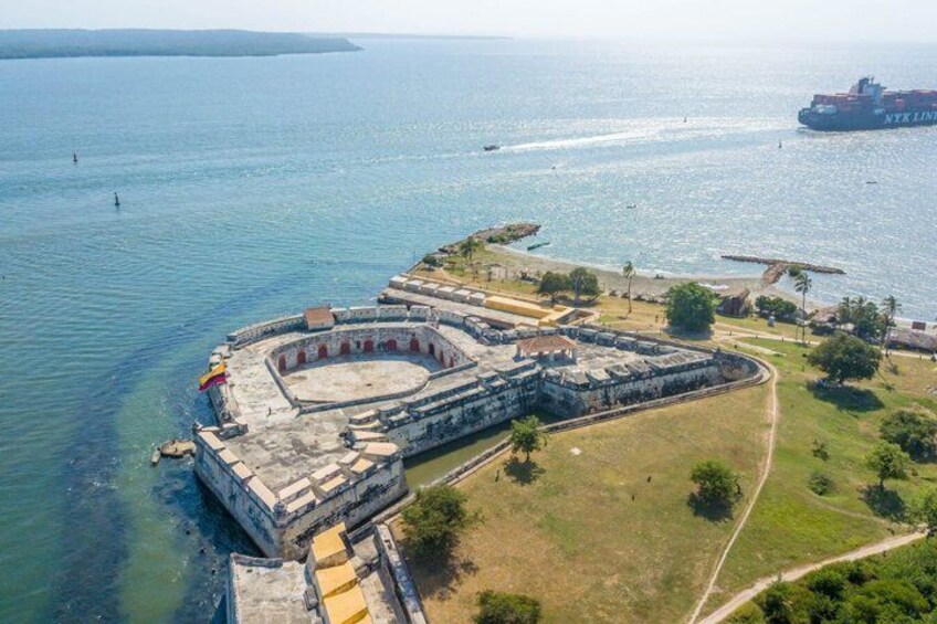 An aerial view of a historical fortification by the sea with a large ship in the background. 1