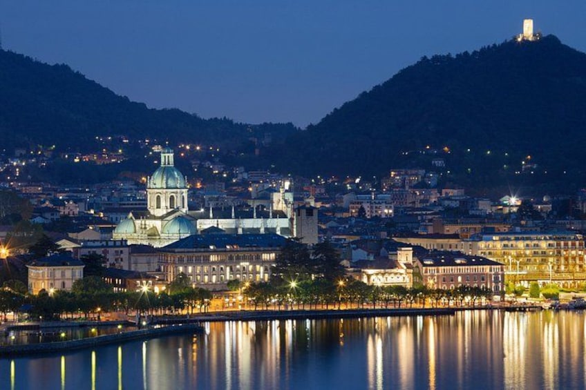 A cityscape at dusk with illuminated buildings, a prominent church with a dome, and a hill in the background. 2