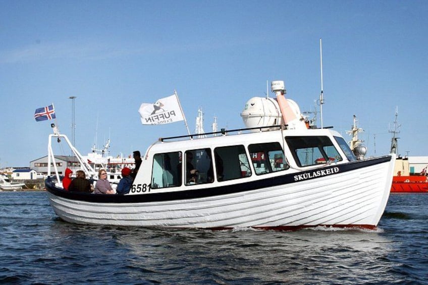A white boat with the name 'SKULASKEID' on the side, a flag, and a person standing at the front. 5