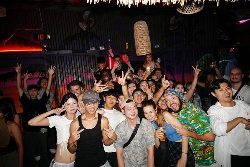 A group of people at a social event, some making hand gestures, in a dimly lit indoor setting with a decorative ceiling fan. 1