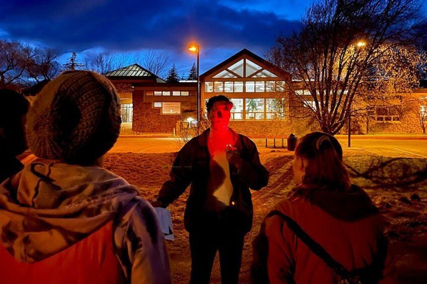 A group of people standing outdoors at night, facing a building with large windows and a triangular roof. 2