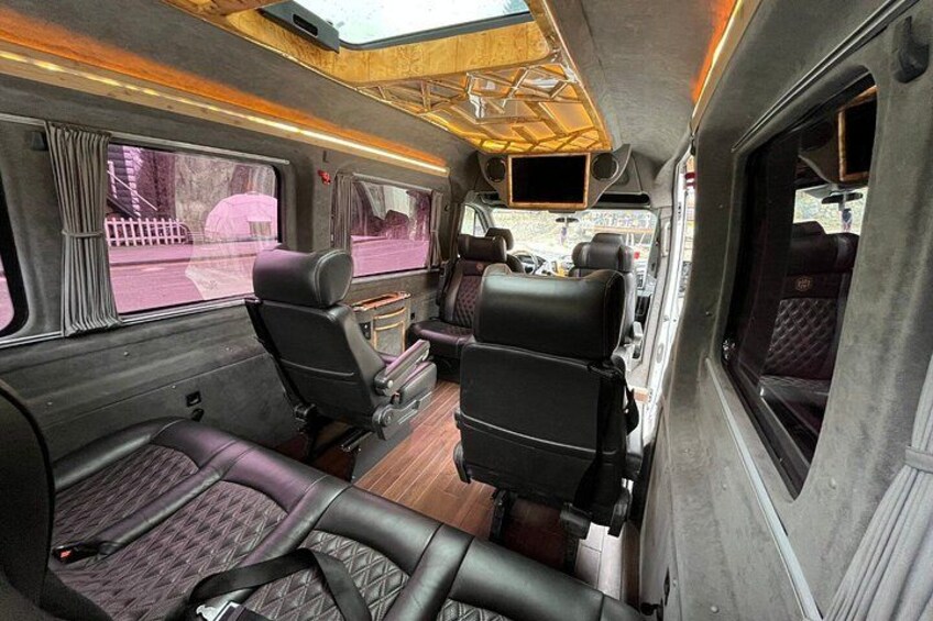 A train interior with wooden flooring, black leather seats, and a window with a view of the outside. 2