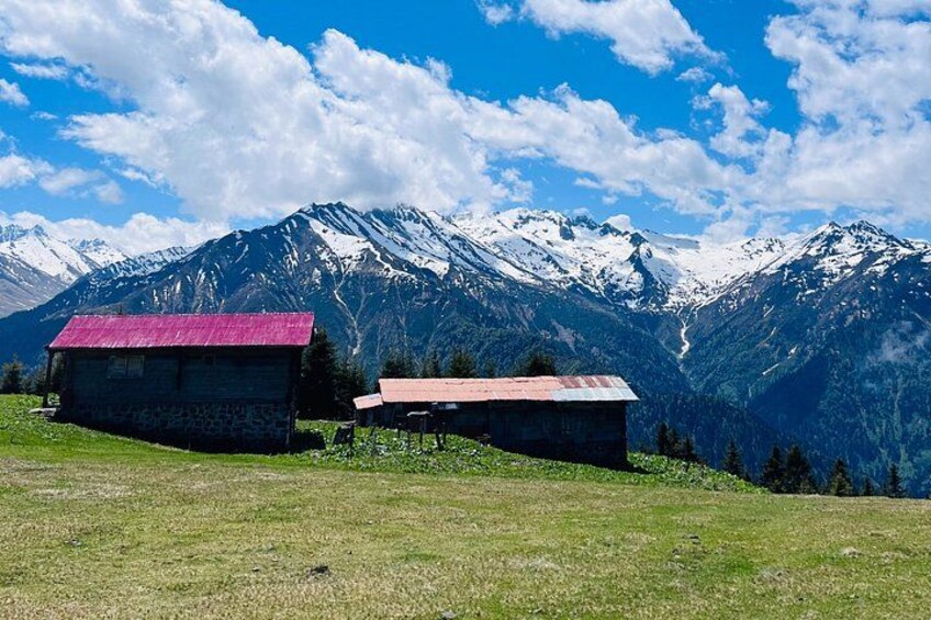 A mountainous landscape with two stone buildings, one with a red roof and another with a brown roof, set against a backdrop of snow-capped peaks under a clear sky. 5
