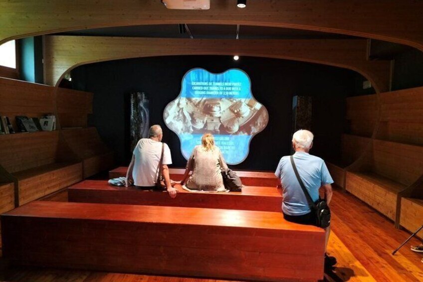 Three people seated on a wooden bench in a museum, facing a large digital display. 2