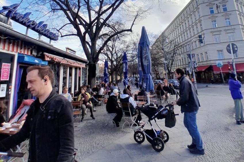 Straßenszene mit Personen, die an Außentischen sitzen, einem Kinderwagen und einem Gebäude mit dem Schriftzug 'Akademie'. 3
