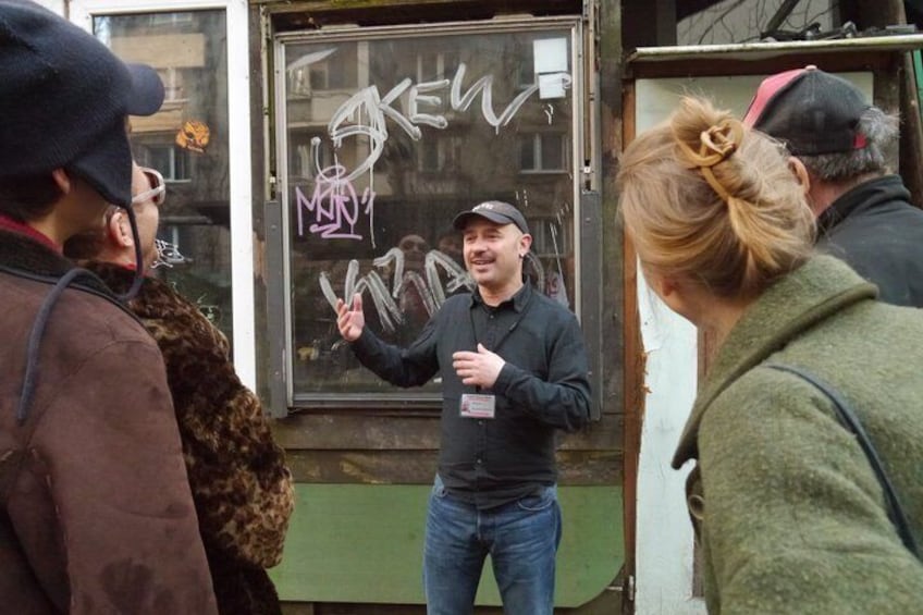 Ein Mann spricht vor einem mit Graffiti beschrifteten Fenster zu einer Gruppe von Menschen. 1
