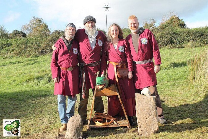 Four people in red coats standing in a grassy field with a windmill in the background. 1