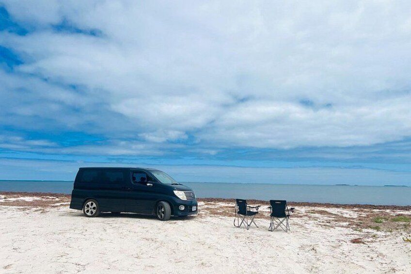 A dark-colored van parked on a sandy beach with two chairs and a clear sky. 3