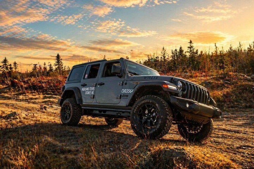 A black Jeep Wrangler with off-road tires and a 'Coyote Tours' decal parked on a grassy field with trees in the background. 1