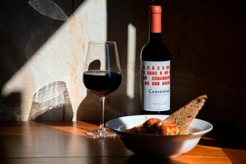 A glass of red wine, a bottle labeled 'Conventud,' and a bowl of food on a wooden table with sunlight casting shadows. 5