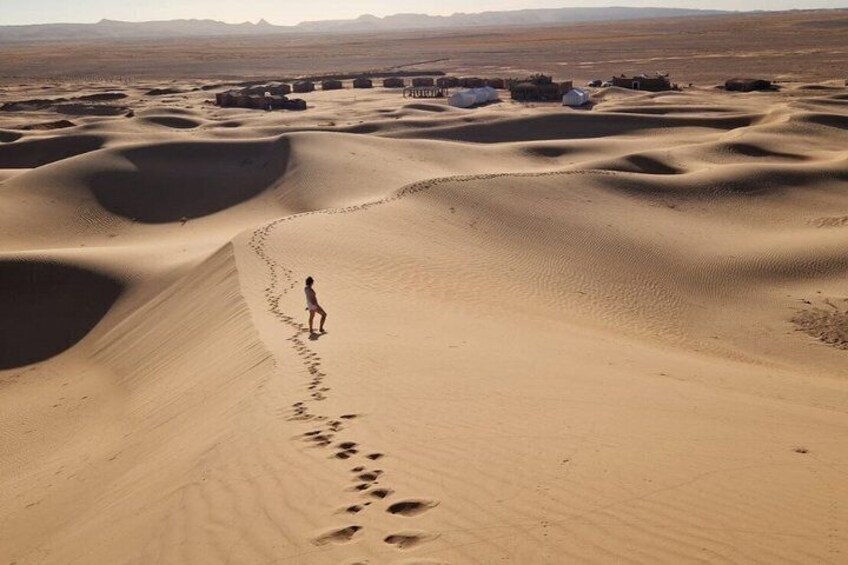 A person walking on a sandy desert with dunes and a few structures in the background. 1