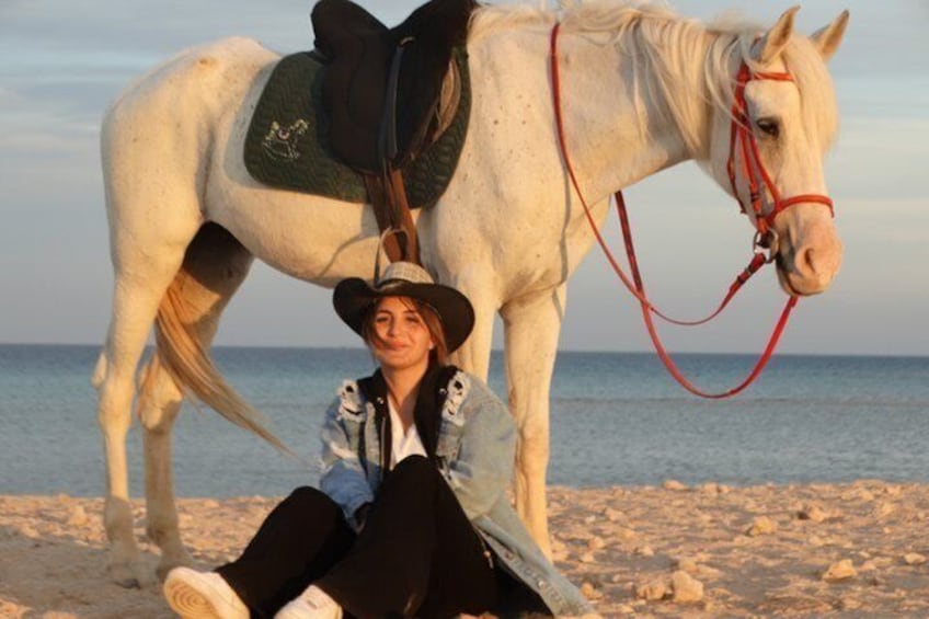 Una persona sentada en la playa junto a un caballo blanco con arnés. 2
