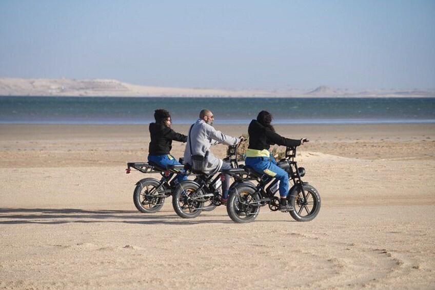 Three people on a tandem bike on a beach with a desert landscape in the background. 3
