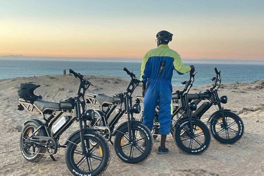 En person står på en sandstrand med flere elektriske cykler parkeret i række. 1