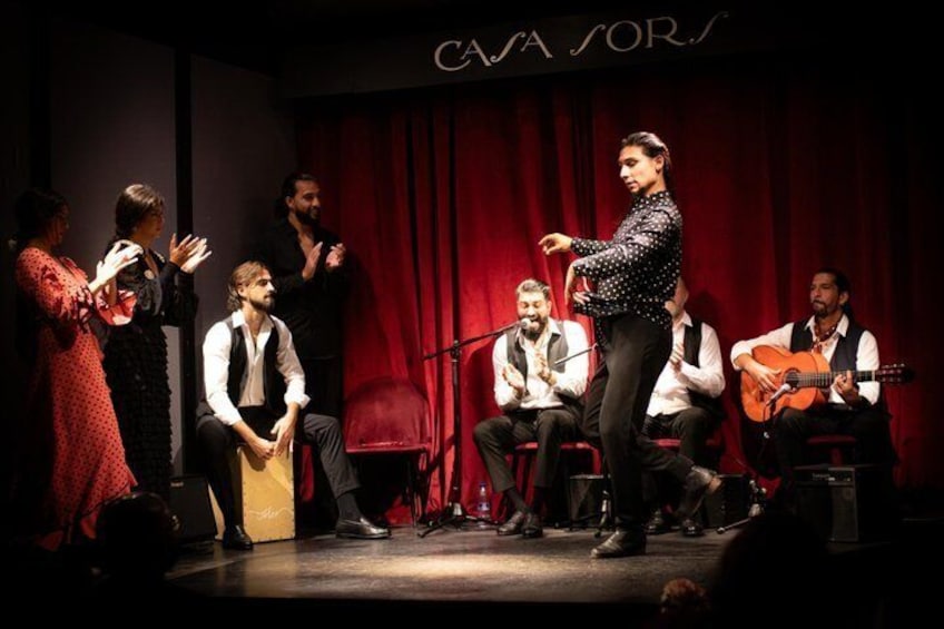 Une scène où se trouve un groupe de personnes, dont certaines jouent d’instruments de musique, avec un rideau rouge en arrière-plan. 4