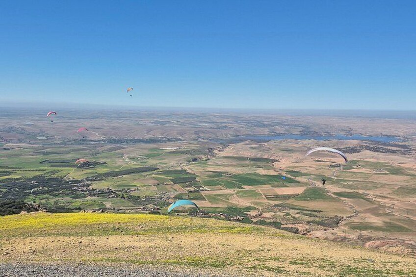 A vast landscape with fields, a small body of water, and multiple paragliders in the distance. 1