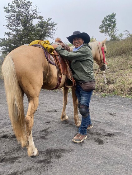 A person in a green jacket and cowboy hat interacting with a light brown horse on a dirt path. 5