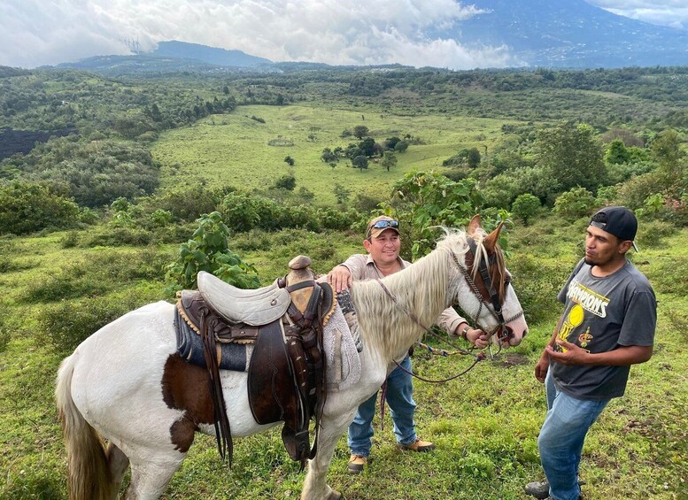 A horse with a saddle and bridle, two people standing beside it, and a lush green landscape with hills in the background. 3