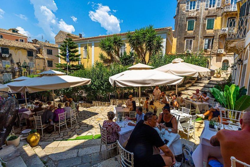 Un café al aire libre con mesas y sombrillas, rodeado de edificios y follaje. 3