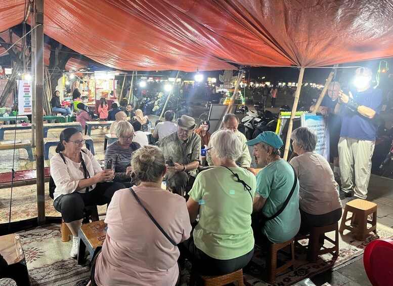 A group of people sitting under a red canopy at an outdoor market. 2