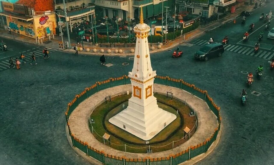 A white monument with a golden spire, surrounded by a green fence, in an urban setting with vehicles and pedestrians. 5
