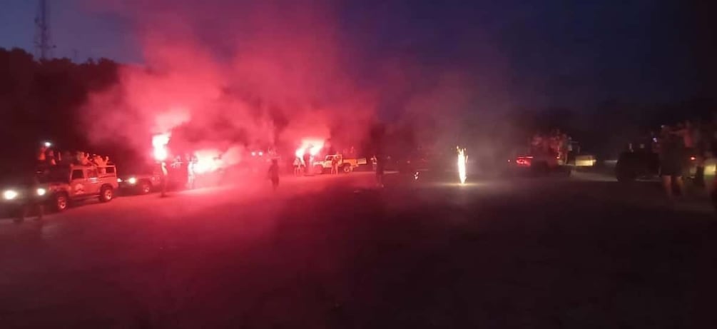 A nighttime scene with vehicles and people, some with lights on, and smoke or fog in the air. 1