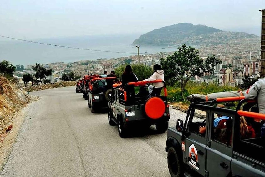 A convoy of off-road vehicles with passengers, traveling on a winding road with a cityscape and mountains in the background. 1