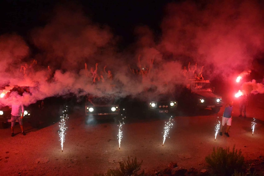 A nighttime outdoor event with vehicles and people, illuminated by red lights and smoke. 3