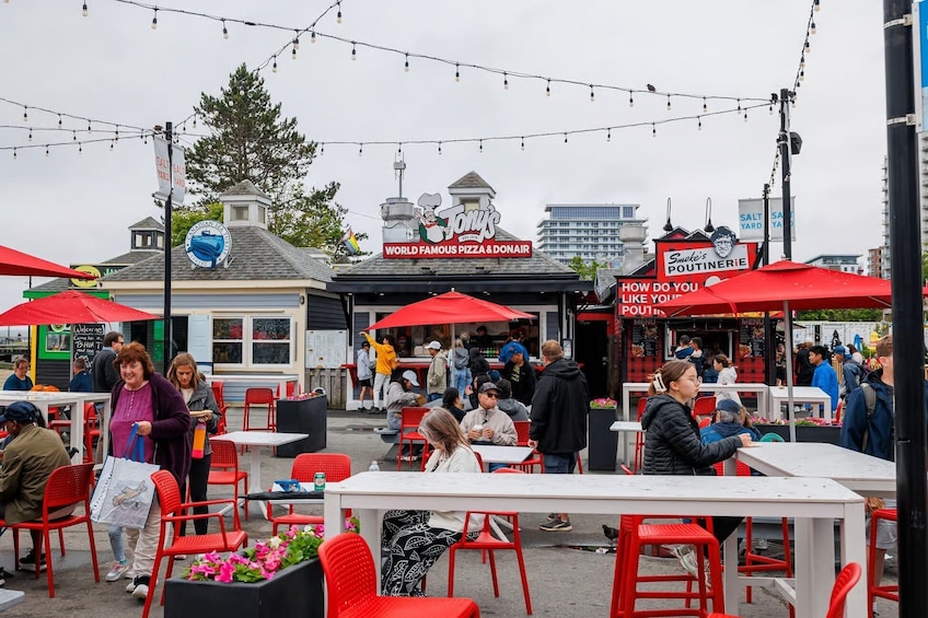 Sitzgelegenheiten im Freien mit roten Stühlen und Tischen, ein Imbissstand mit dem Schild 'WORLD FAMOUS PIZZA & DONAIR' und ein Gebäude mit dem Schild 'SALTY YARD'. 2