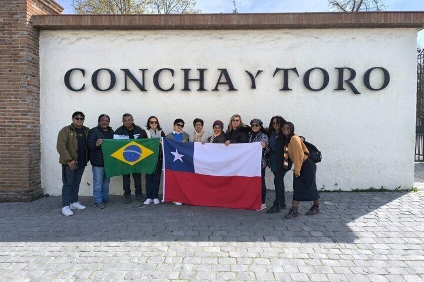 A group of people standing in front of a wall with 'CONCHAYTORO' written on it, holding a Brazilian flag and a flag with a white, red, and blue design. 1