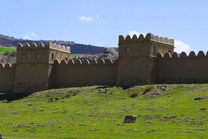 A medieval-style stone wall with crenellations, a grassy field, and a hilly landscape in the background. 2