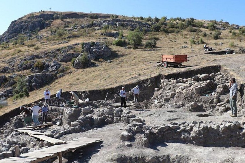 An archaeological excavation site with people working on the ground, a wooden structure, and a red vehicle. 3