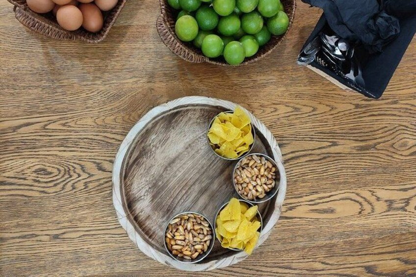 Een houten tafel met een rond dienblad waarop gele bloemen, pijnboompitten en een schaal groene limoenen liggen. 3