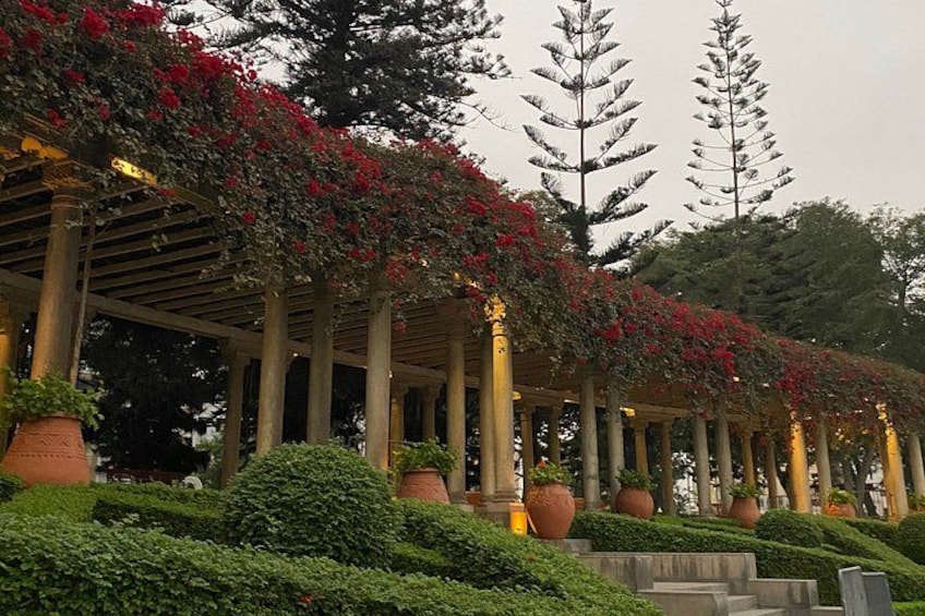 A covered walkway with columns, red flowers, and potted plants. 1