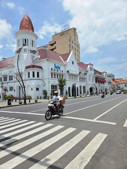 A street with a white building with a red roof and a tower, a motorcyclist with a delivery box, and a clear sky. 4