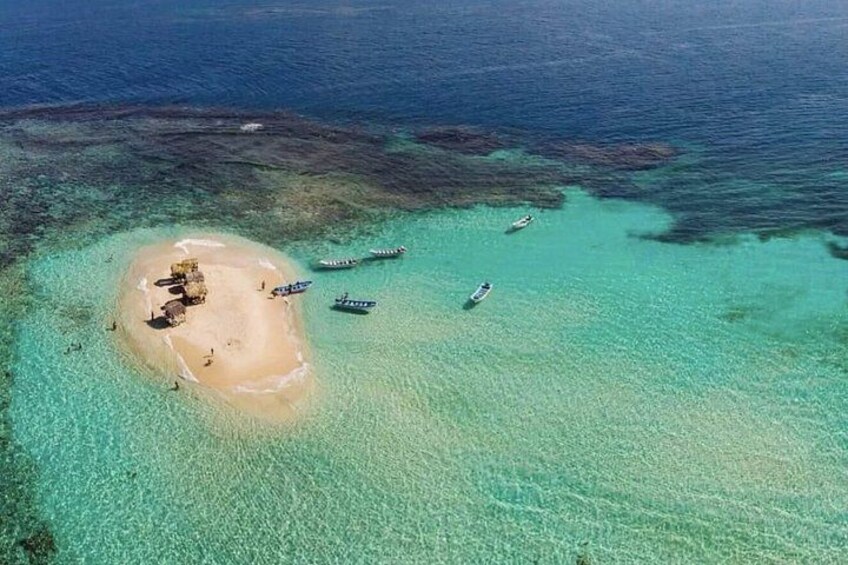 Ett flygbildsbild av en liten ö med kristallklart turkost vatten, en sandstrand och flera båtar som ligger ankrade nära stranden. 5