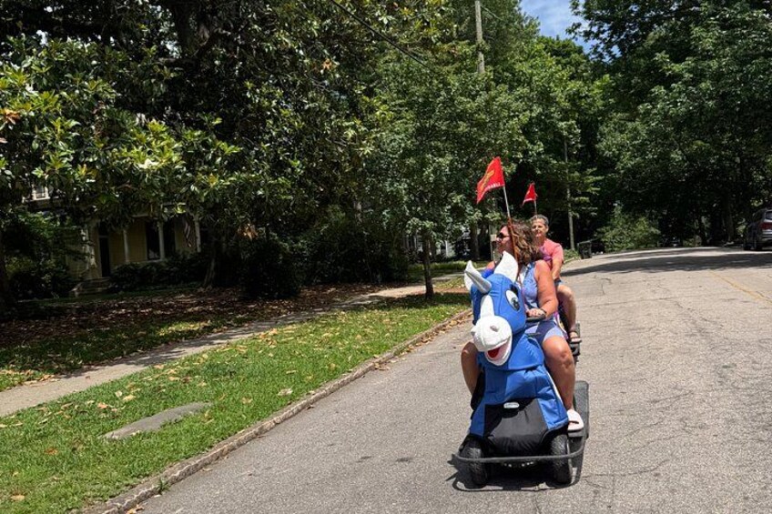 Two people riding a blue horse-shaped scooter down a residential street. 4
